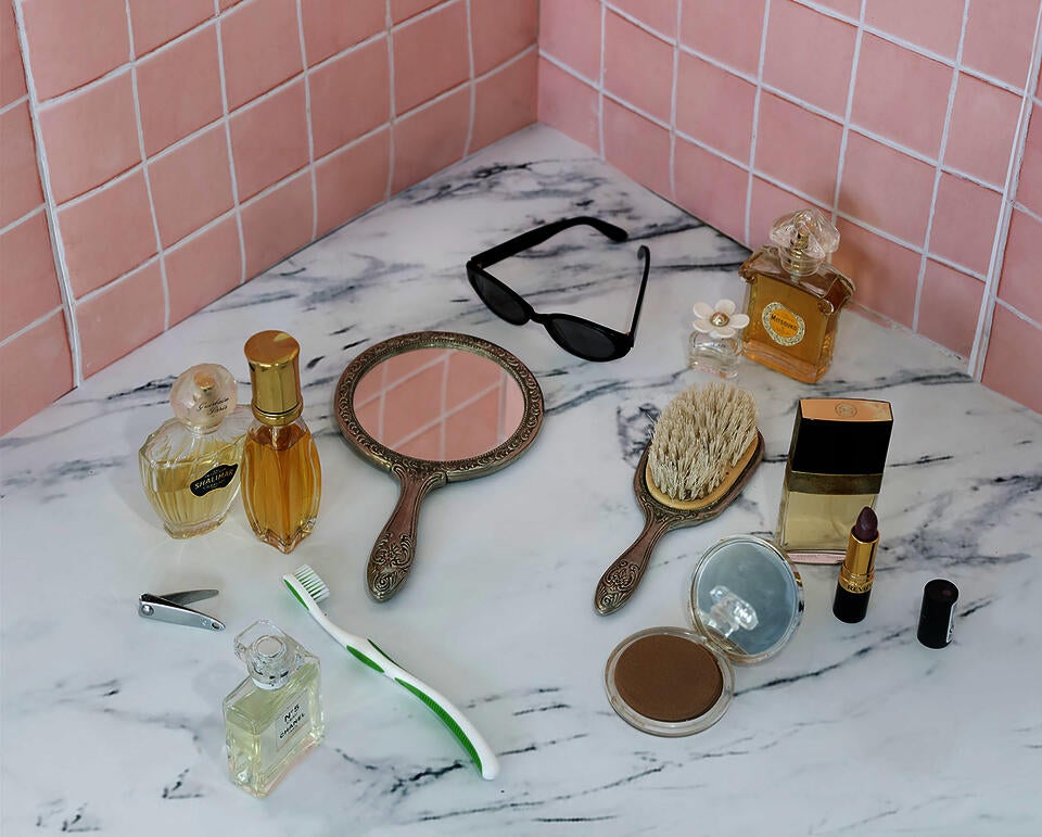 A still life of a marble countertop against pink ceramic tiles with perfume bottles, a hairbrush, compact mirror, sunglasses, and makeup.