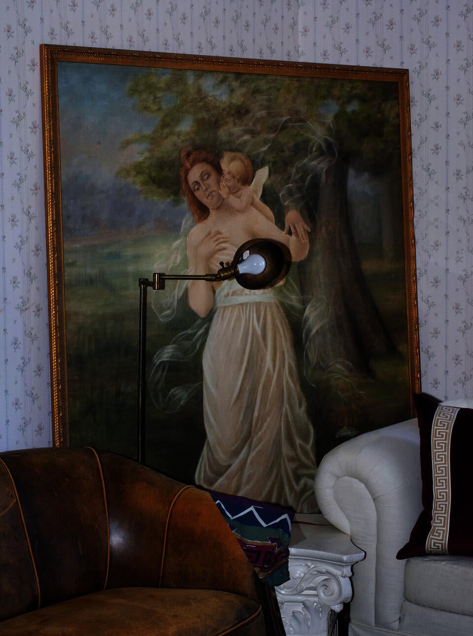 A brightly-lit image of a large painting of Venus and Cupid flanked by two lounge chairs and a column-like pedestal. The Venus figure in the painting is obscured by a floorlamp in the middle-ground. 