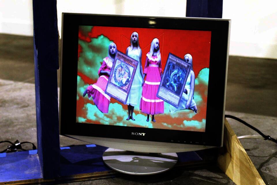 A still from the video on a silver Dell monitor shows the artist four times in pale makeup, wearing a bright pink period dress and a pale blue nightgown, symmetrically on a red and green horizon. A pentagram rotates in the background.