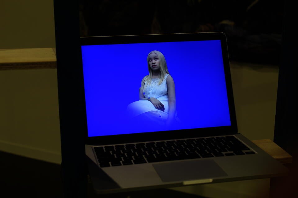 The image shows the artist in a video still, wearing pale face paint, a blond wig, and a pale blue nightgown, against a fluorescent blue background. The video plays on a MacBook Air 2019.