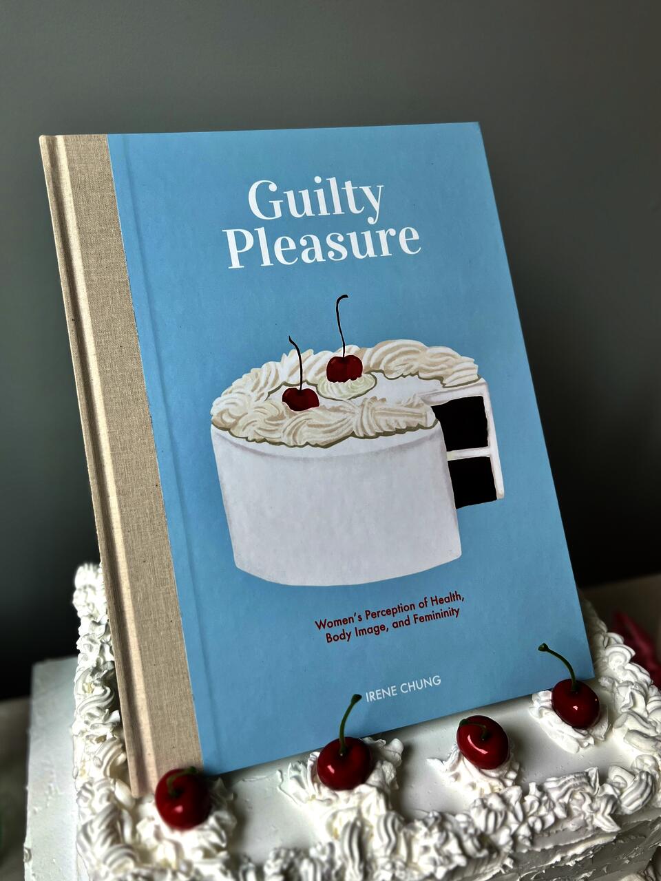 an illustrated book, titled Guilty Pleasure, sitting on a cake with cherries on top 