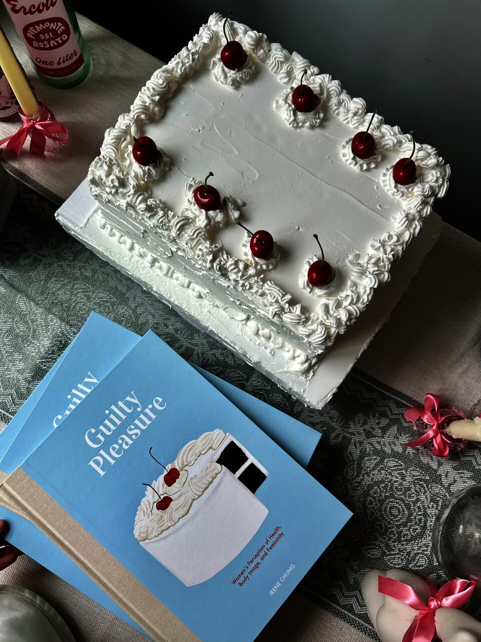 photo of illustrated books next to a white-cream cake with cherries on top, on the surface of tablecloth