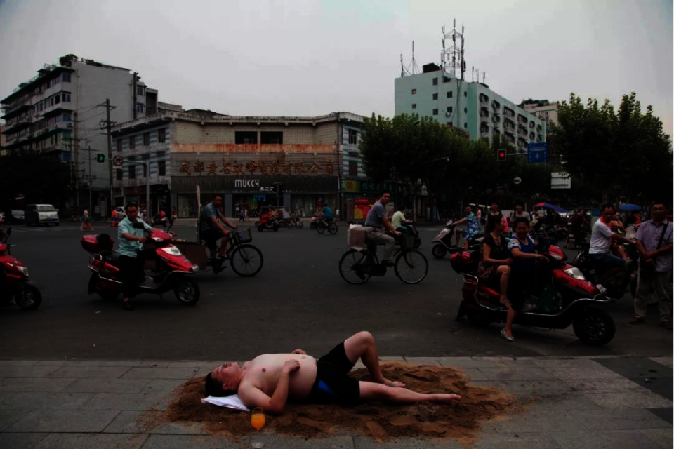 The artist lying on a mat on a tiny beach he made in a public area with only shorts. He's on a vacation.