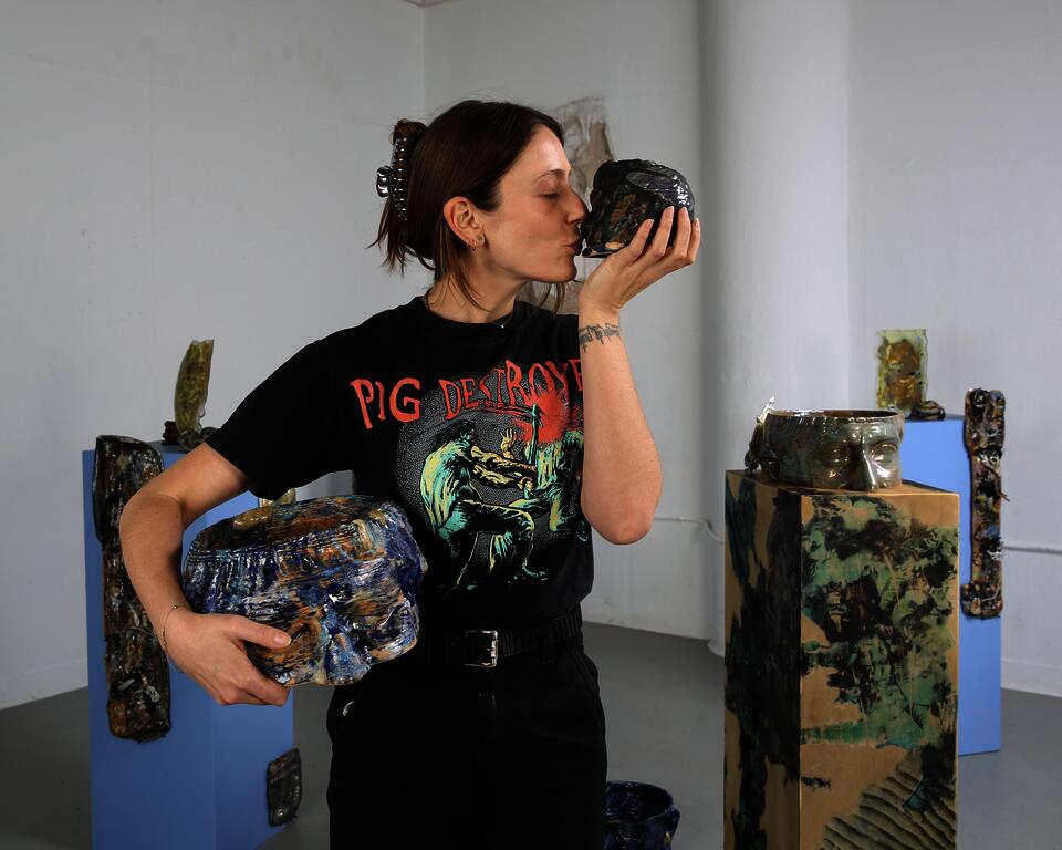 Portrait of artist standing in front of her installation. Turned to her left and kissing a small ceramic 3D printed head.   