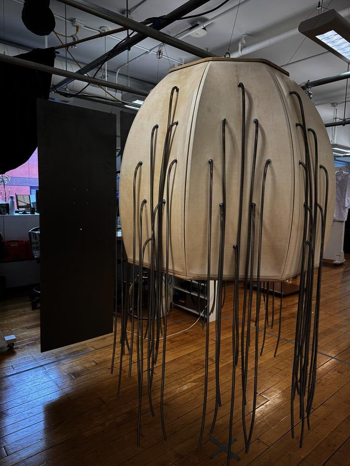 Barrel-shaped goatskin structure with hanging metal springs, surrounded by steel sheets and lit by overhead projection.