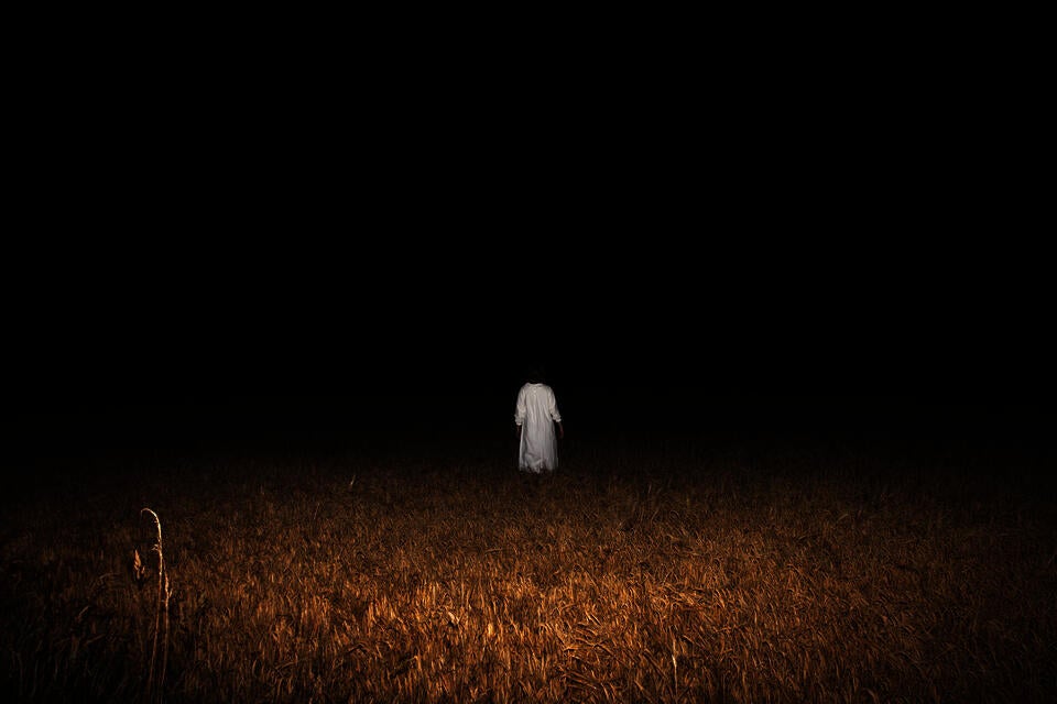 A figure in a white Sankta Lucia gown stands in a wheat field. She faces the dark, void of the night.