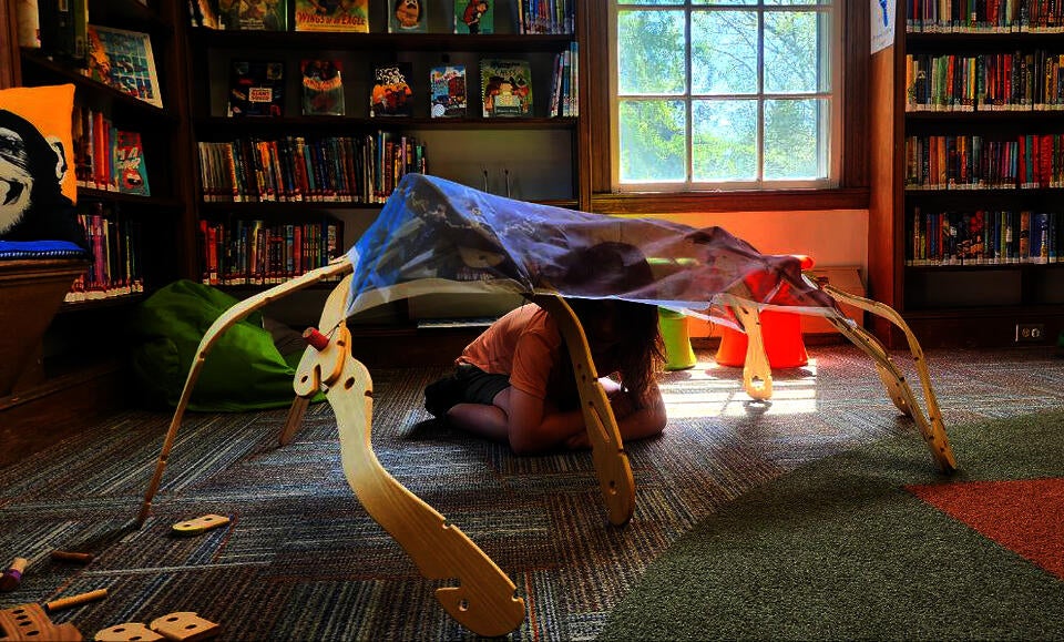 Child sitting inside a wooden toy structure covered in printed fabric with ash tree leaves, seeds, bark, and twigs