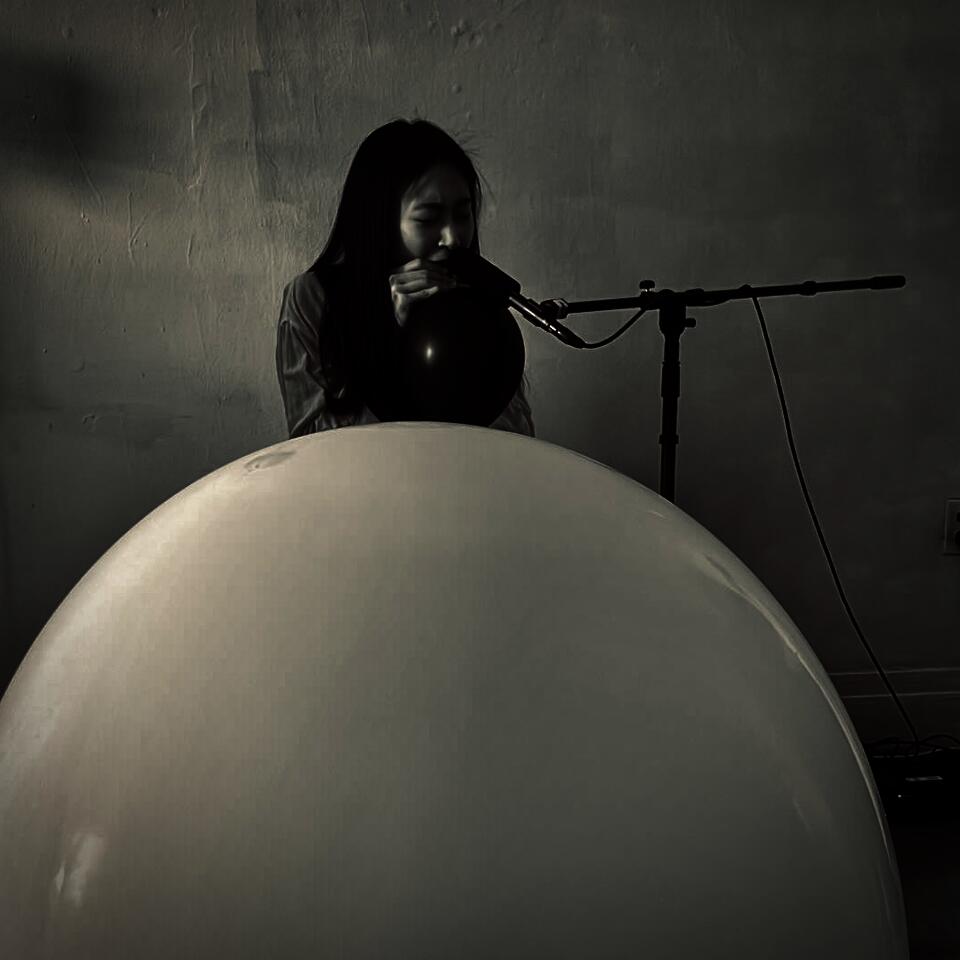 A performer creates balloon noise with a microphone, processed through a computer, and played back via speakers.