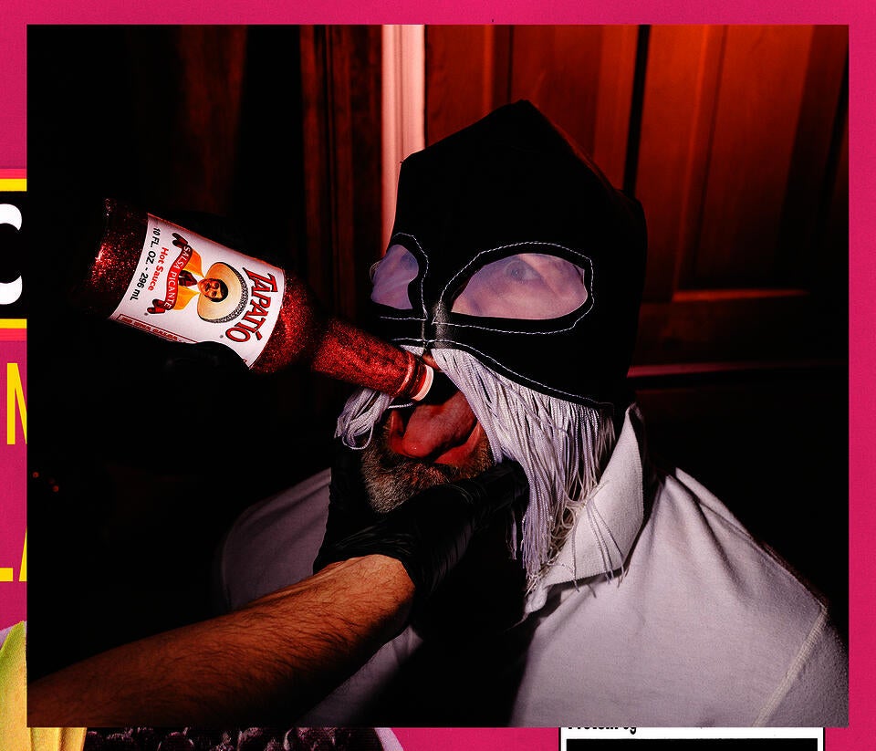 A masked man in a lucha libre hood with white fringe is force fed pink glittered Tapatío on his tongue by an unknown hand