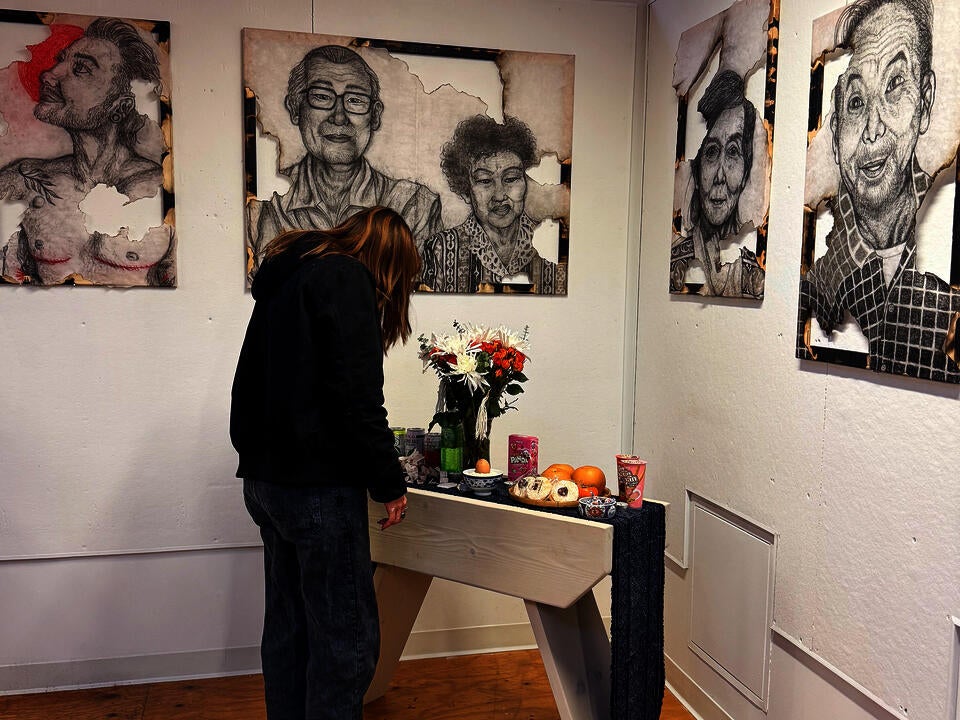 A person stands and examines an altar holding food and fresh flowers. On the walls are partially burned charcoal portraits.