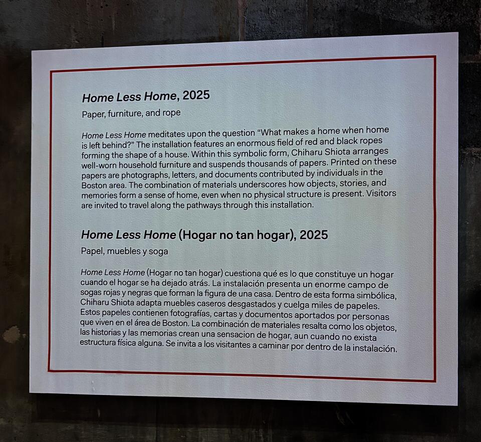 A large white label affixed to a concrete wall. The name of the work, the materials used, and a brief paragraph about the installation written in English; below, the name of the work, the materials used, and a brief paragraph about the installation written in Spanish. 