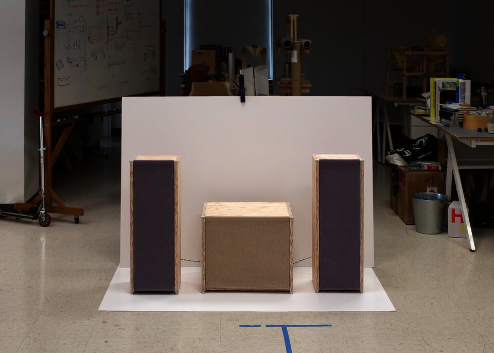 Two pale plywood speakers with hanging fabric fronts symmetrically flank a lower cabinet with a similar fabric front in a design studio, set against a large white backdrop with sketches, tools, and prototype furniture visible in the background.