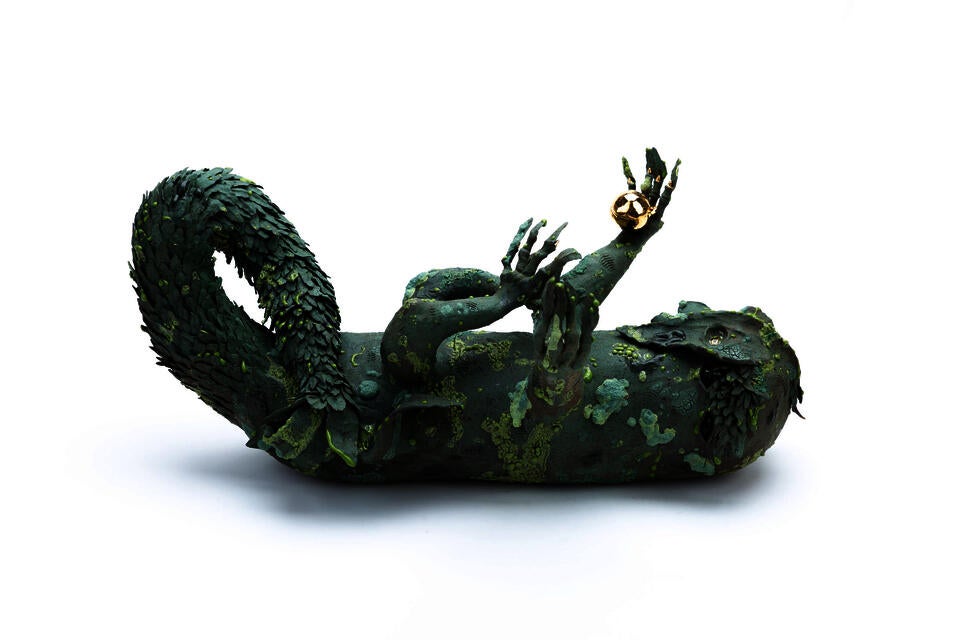 A green four legged creature lying on its back and reaching up to the sky. It has a twisting leafy tail, and is covered in mossy texturing. It holds a golden bell in one hand.