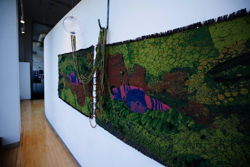 Jacquard-woven textile with lying figure among green forms, with draped glass beads connecting to a hanging glass orb