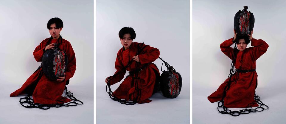 A model wearing a red traditional Chinese Tang dynasty robe holds an oversized wine vessel sculpture against the body or lifts it above the head. Black braided hair forms loop around the figure and gradually increase in size from necklace-scale rings to large circular forms surrounding the vessel.