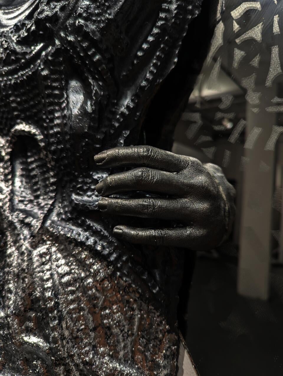Detail chain-textured cement form, partially silver over gray, surrounded by mirrors, faintly etched with Hmong batik patterns. A silver-gray hand emerges from one mirror, reaching toward the cement in an ambiguous gesture of embrace or pull.