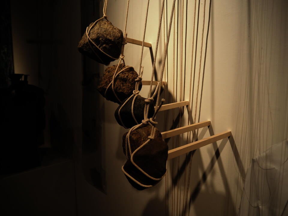 Five rocks suspended by sticks compress five wood sticks against the wall, holding them in the air.