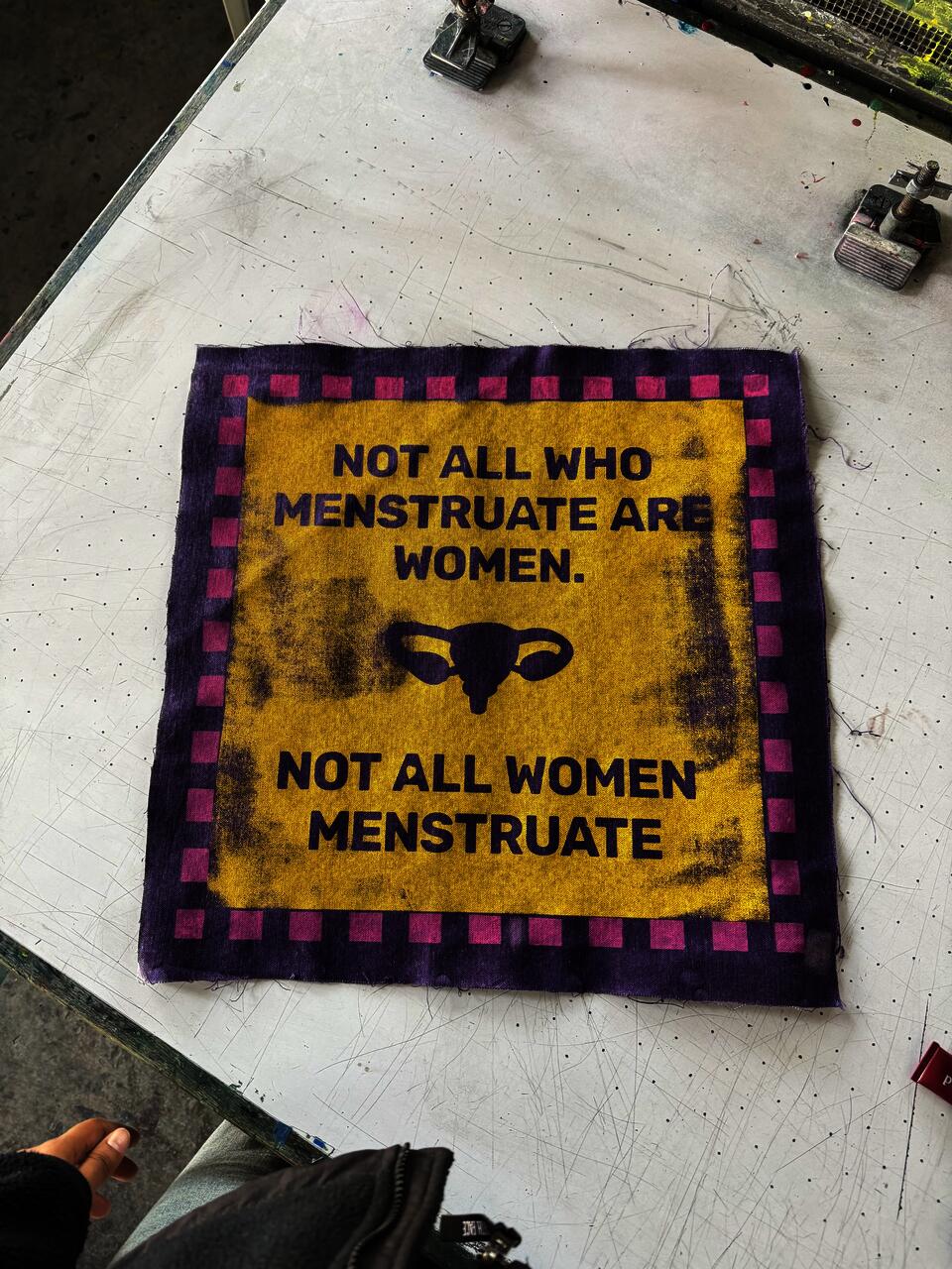 "Screen print on fabric, yellow, purple & pink A bold, graphic patch printed in purple on a golden-yellow field with a pink-and-purple checkered border. The text declares: ""Not All Who Menstruate Are Women. Not All Women Menstruate."" — flanked by a uterus icon. Photographed fresh off the print table."