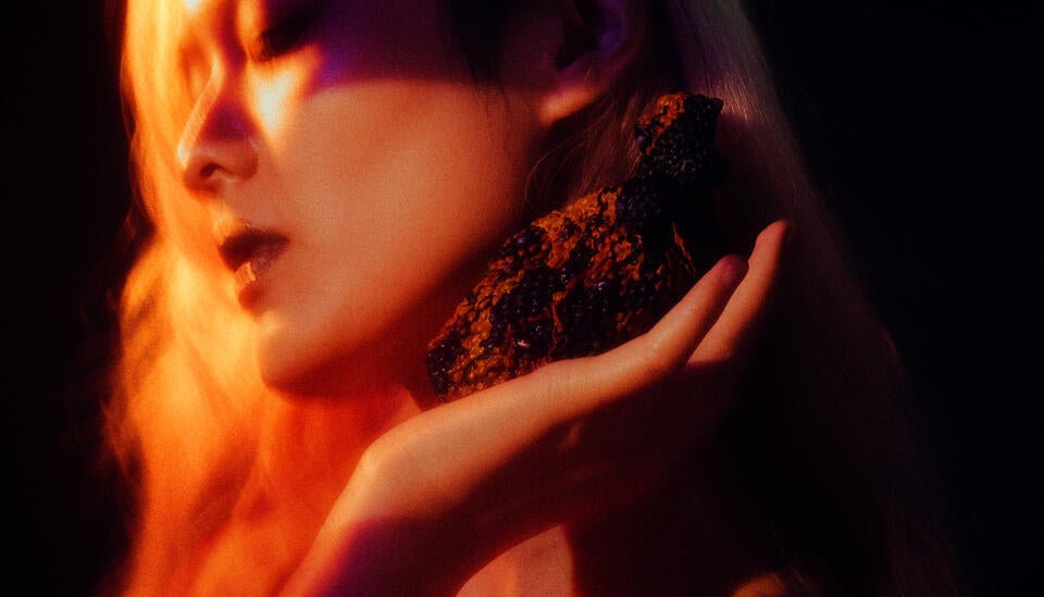 Textured orange-brown heart-like object rests in a hand at right, raised to the ear; clustered dark nodules catch warm amber light, fading into deep purple shadow, creating an intimate, tactile focus.