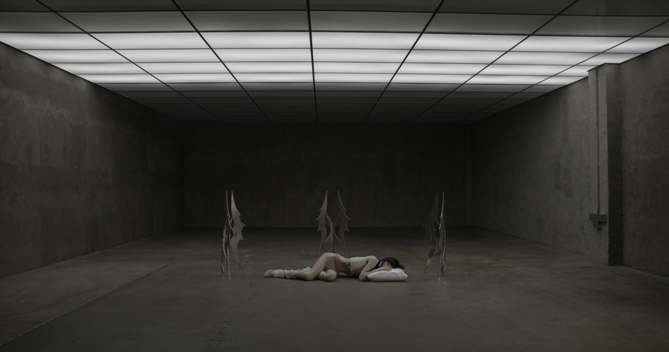 Wide shot of person lying on floor within large empty gray room, surrounded by metal sculptures, head resting on white pillow under grid ceiling lights.