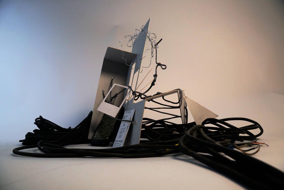 Concept model consisting of white panels, wire, and rope entangled with thick black cables, with an iphone displaying a map of Gaza, arranged against a white background.