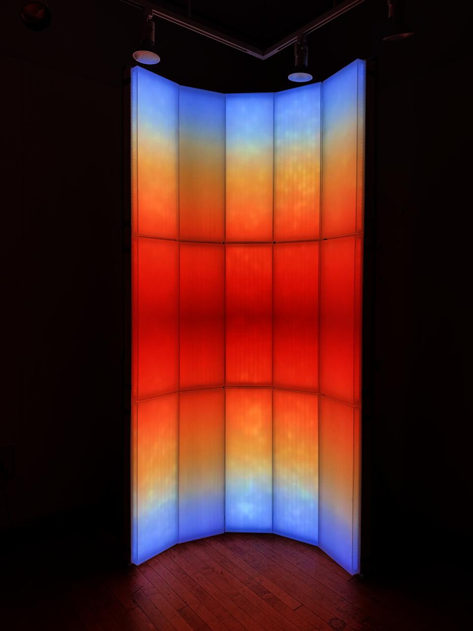 A light-based installation composed of vertical translucent panels, with blue, orange, and red gradients that evoke the color transition of sunrise and sunset.
