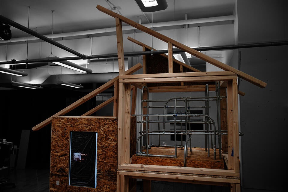 A close-up view of the architectural installation frame, constructed from raw lumber. On the left side, a section is enclosed by a textured particleboard (OSB) wall. 
