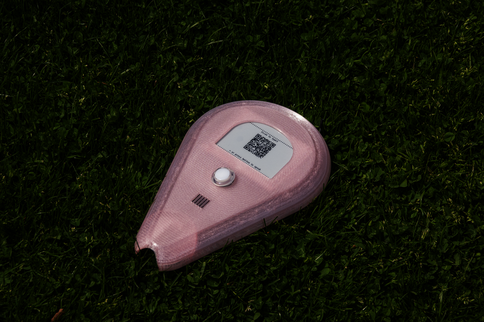 Pink teardrop-shaped interactive device resting on grass, featuring a button, speaker, and QR code displayed on a screen.