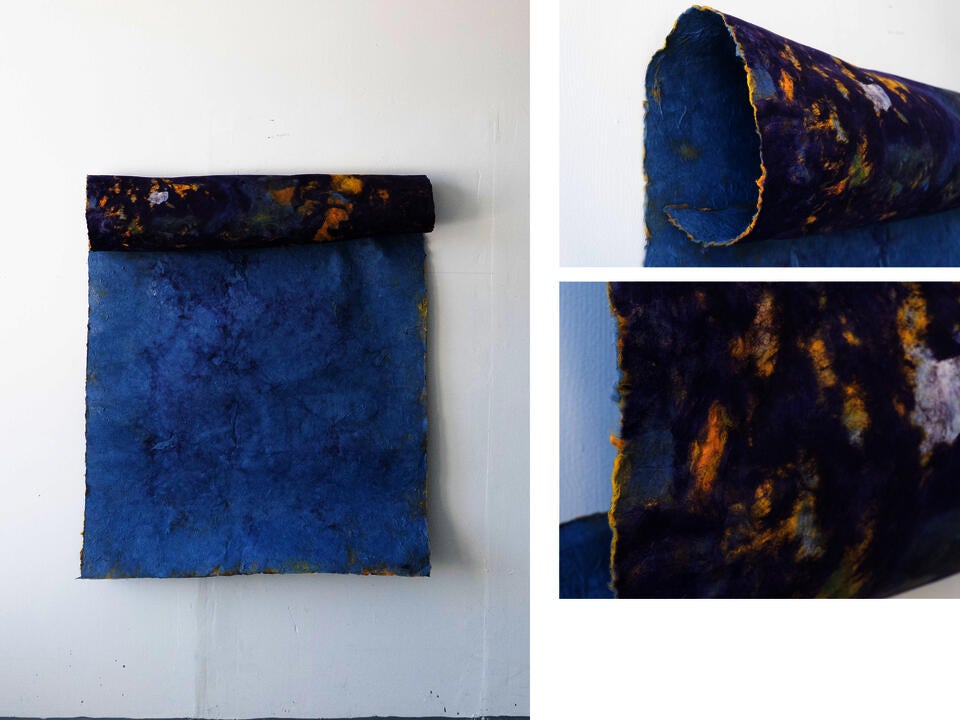 A large sheet of handmade blue paper hangs from a rolled cylindrical form attached along its top edge. The cylinder is covered in marbled purple, yellow, white, and blue textures. Close-up images show the rough edges