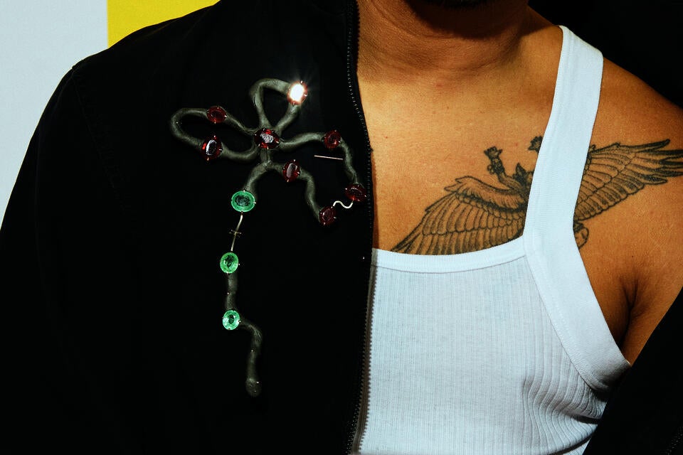 A brooch loosely in the shape of a flower with three large petals, made up of grey epoxy and metal wire. Rubies and sapphires are set in prong settings along its length. Worn by a man with a vest, closely cropped to his chest.