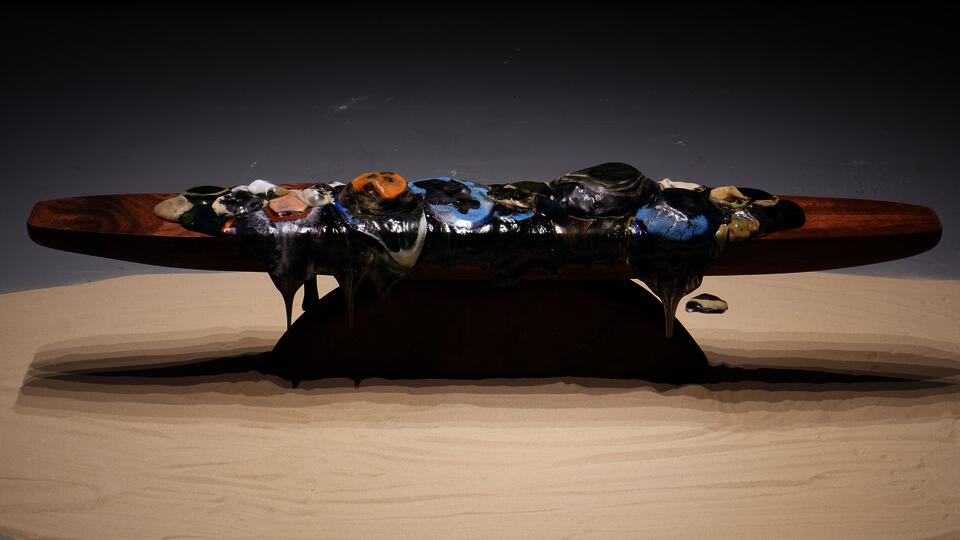 Molten glass forms in varied colors spread and drip across a long wooden platform, with softened edges and flowing material extending downward.