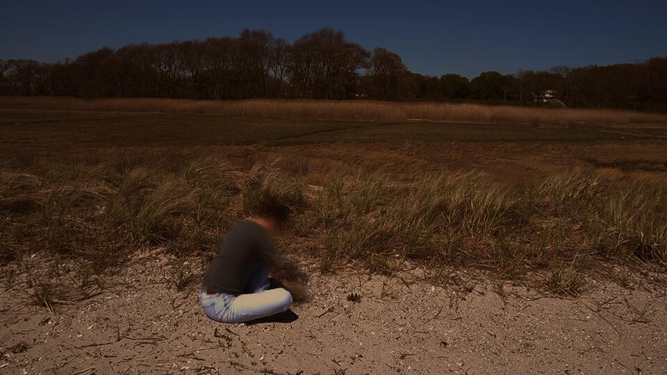 A person sits in a sandy dune landscape while making a bowl-shaped form in the ground. Marsh grasses and trees extend across the background beneath a clear sky.