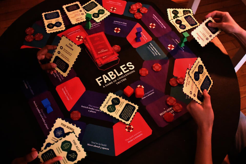 A colorful board game layout displays "Fables: A Game of Challenging Truths" with character archetype cards, scenario cards in red and purple, coral-colored tokens, and red dice, arranged on a wooden table with players' hands holding cards.