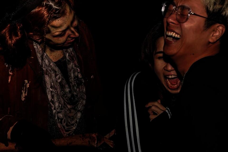 Three people are visible in the dark at a haunted attraction park. One is a scare actor in a scary costume. The other two, visitors to the attraction, huddle together in response to the scare.
