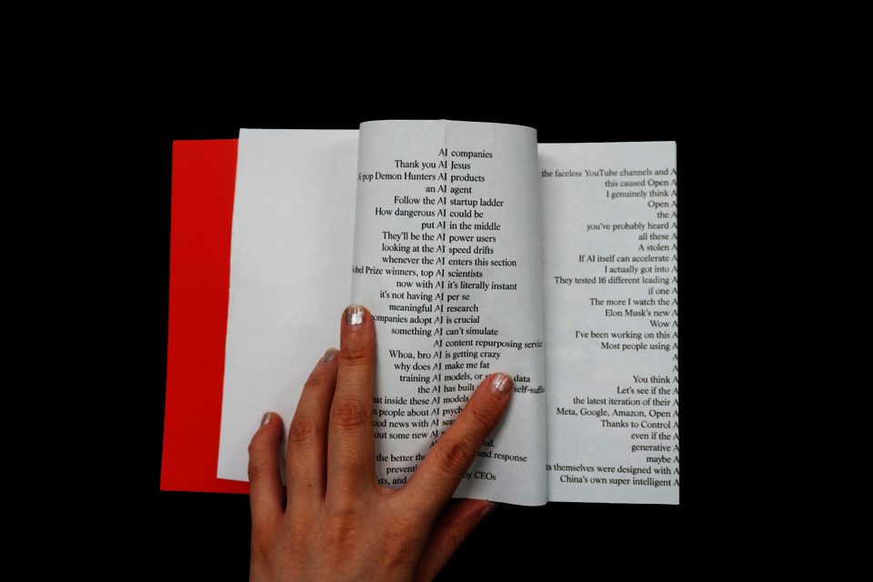 a book with a red cover against a black background. The interior spread shows two columns of text, each line containing a sentence fragment with "AI" as the pivot point — the left half of each sentence ends with "AI" and the right half begins after it, creating a visual caesura down the center of each page. 